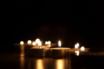 Spirituelle Bildung und Begleitung Das stimmungsvolle Bild zeigt, wie in einem dunklen Raum auf einem Tisch brennende Teelichte stehen, deren Leuchten sich auf der glatten Tischplatte spiegelt